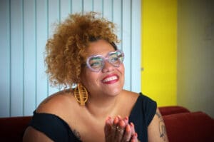 Bianca is a fat light skinned disabled queer AfraLatina who is smiling and looking to the right. Her hands are clasped and we see her orange fingernails and gold rings. She is wearing light blue heart shaped glasses, red lipstick, and has large round gold earrings. Her hair is up and her blondish brown curls cascade around her head. A black t-shirt that scoops across her shoulders reveals her many tattoos on her arms.