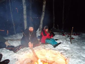 2 people sitting around a fire in snow, one of them has a sick in the fire.